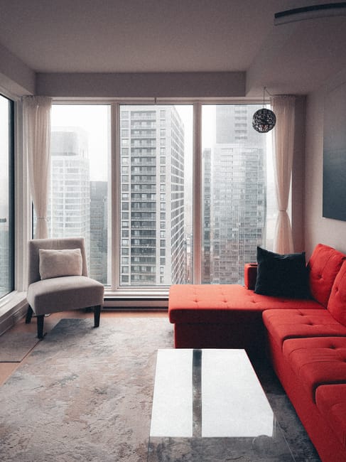 A bright, modern living room with a large light gray fabric sofa adorned with black, white, and gray cushions, positioned against a white wall. To the left, a tall floor lamp with a round white shade 